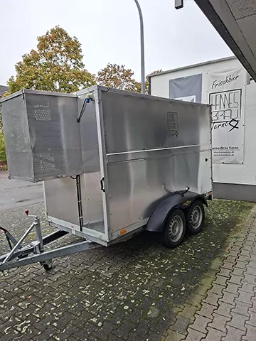 Der Kühl-Anhänger: geschlossener Aluminium-Anhänger mit Doppelachse und HannesBräu-Logo, vorn links der Aufbau mit Lochblech